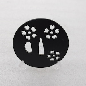 Op de afbeelding: Zwarte ronde metalen tsuba met een decoratief ontwerp van uitgesneden vijfbladige bloemen. De tsuba is een traditionele Japanse zwaardbeschermer.
