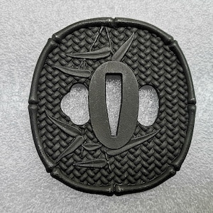 Op de afbeelding: Zwarte metalen tsuba, een decoratieve bescherming voor een Japans zwaard, met een geweven patroon en een gestileerd bamboe ontwerp.