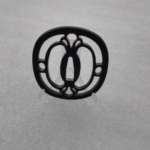 Op de afbeelding: Zwarte metalen tsuba, een decoratieve bescherming voor een Japans zwaard, met een ronde vorm en een ingewikkeld uitgesneden ontwerp.