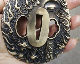 May include: A circular, brass-coloured sword guard with a detailed design of flames and a stylised face. The guard has a central opening for the sword blade and features a swastika symbol and Japanese characters.