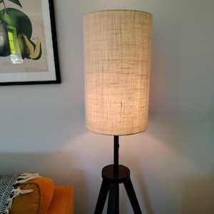May include: A floor lamp with a beige linen shade and a black tripod base. The lamp is switched on and casting a warm glow.
