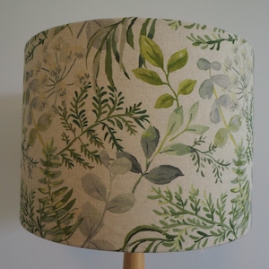 May include: A beige lampshade with a green and grey leafy pattern. The lampshade is cylindrical and has a smooth surface.