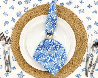 May include: A white dinner plate with a blue and white floral patterned napkin on top of it. The plate is on a brown woven placemat. There are two forks and two spoons on the white tablecloth with a blue and white floral pattern.