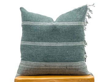 May include: A teal blue and white striped throw pillow with fringe on the edges sits on a wooden stool.