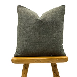 May include: A square, dark gray decorative pillow with a textured fabric. The pillow is placed on a small, light brown wooden stool. The pillow has a simple, neutral design, suitable for various home decor styles.
