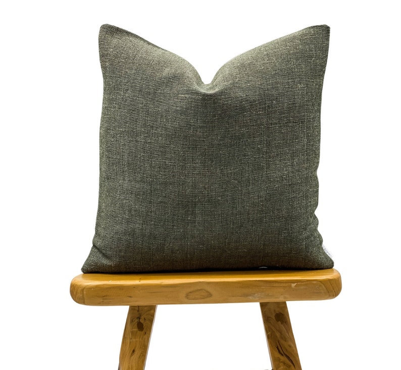 May include: A square, dark gray decorative pillow with a textured fabric. The pillow is placed on a small, light brown wooden stool. The pillow has a simple, neutral design, suitable for various home decor styles.