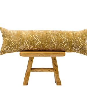 May include: A long, mustard yellow lumbar pillow with a white polka dot pattern. The pillow is sitting on a wooden stool.