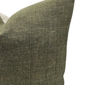 May include: Close-up of a textured, olive green pillow. The fabric has a woven appearance with a mix of dark and light threads. The pillow's corner is visible against a white background.