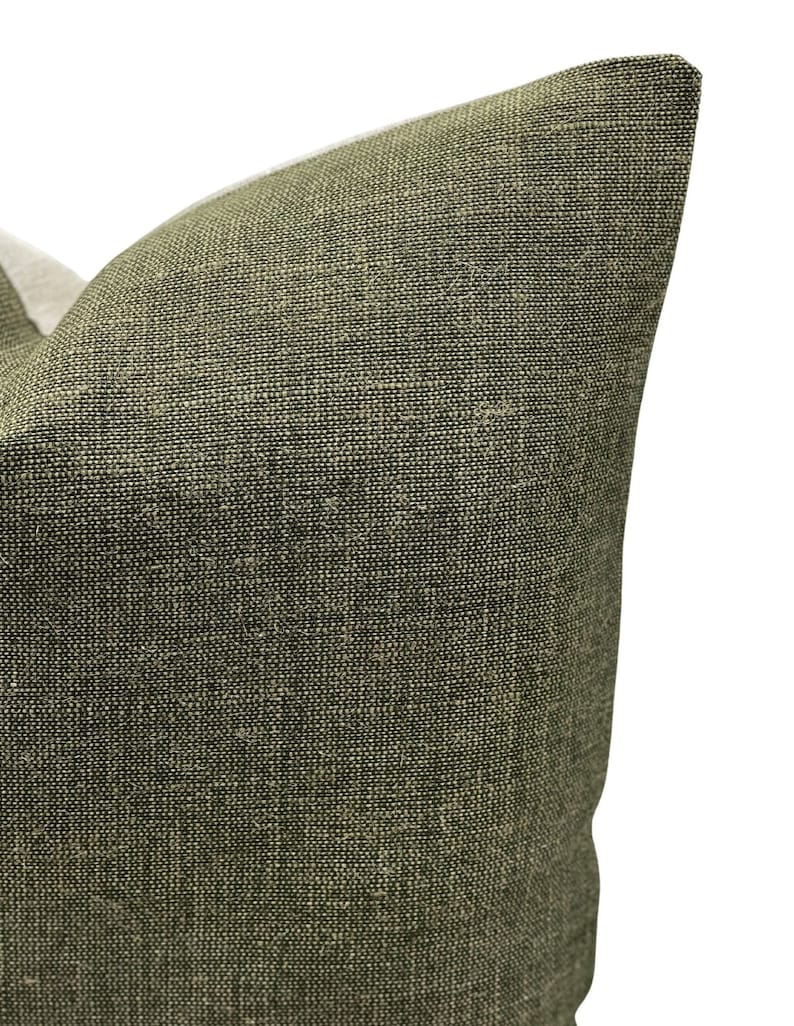 May include: Close-up of a textured, olive green pillow. The fabric has a woven appearance with a mix of dark and light threads. The pillow's corner is visible against a white background.
