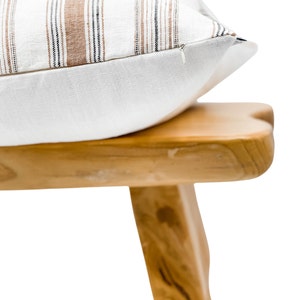 May include: A white pillow with brown and black stripes sits on a wooden stool. The pillow has a zipper closure.
