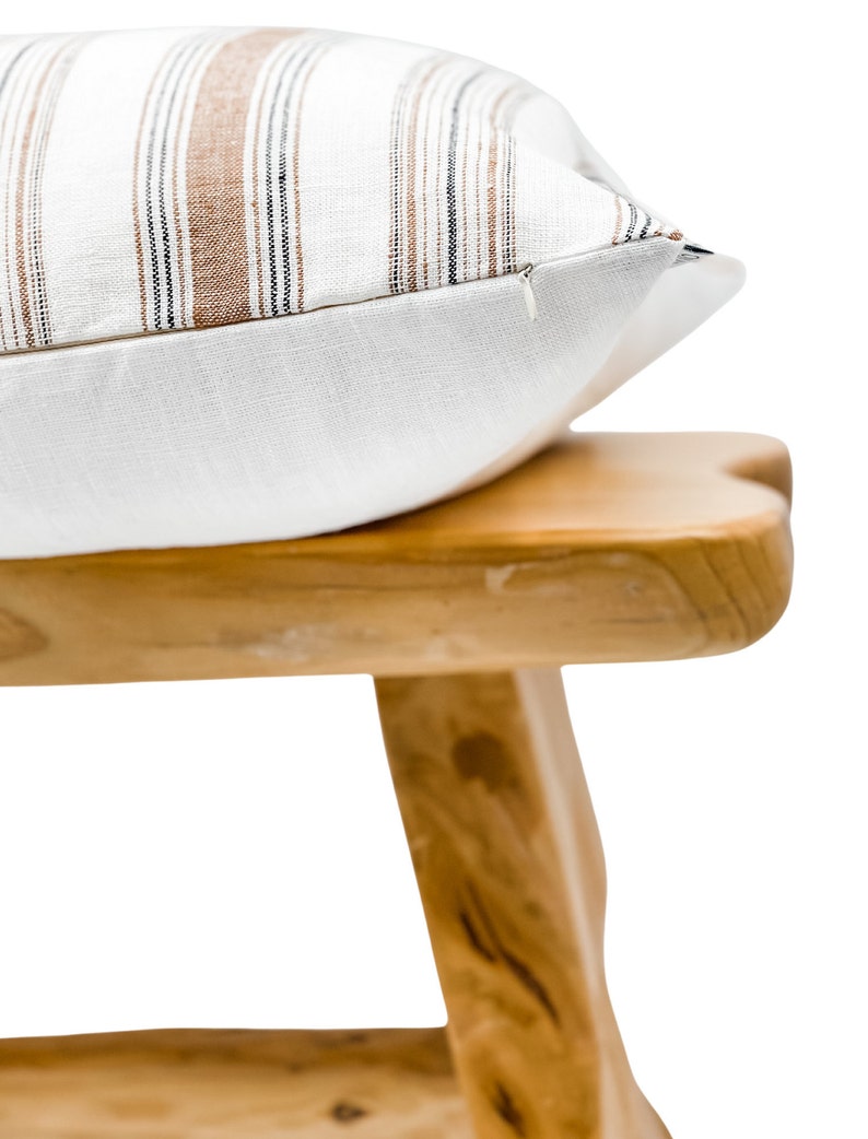 May include: A white pillow with brown and black stripes sits on a wooden stool. The pillow has a zipper closure.