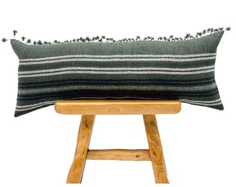 Op de afbeelding: Een lang, rechthoekig sierkussen met een donkergroene achtergrond en horizontale strepen in wit en zwart. Het kussen heeft kwastjes langs de bovenrand en wordt gepresenteerd op een houten kruk. Woondecoratie-artikel.