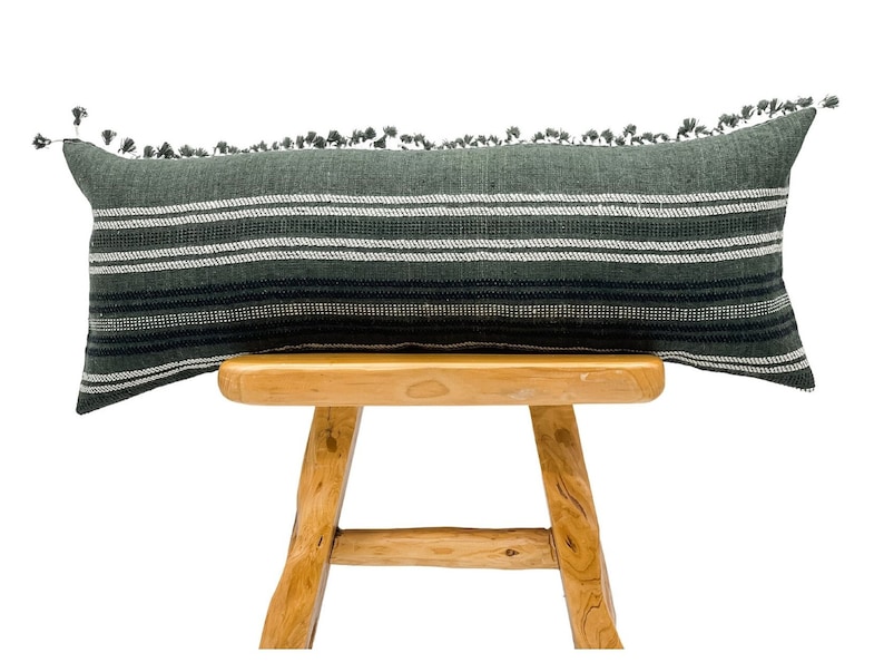 Pode incluir: Uma almofada decorativa longa e retangular com fundo verde escuro e listras horizontais brancas e pretas. A almofada tem borlas ao longo da borda superior e &eacute; exibida em um banquinho de madeira. Artigo de decora&ccedil;&atilde;o para casa.