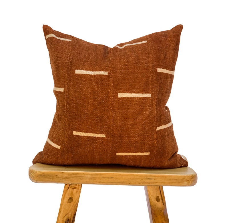 May include: A brown throw pillow with white stripes sits on a wooden stool. The pillow is made of a textured fabric and has a simple, modern design.