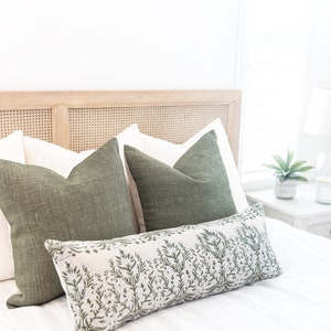 May include: A bed with a light wood headboard and white bedding. Two green square pillows and a long, rectangular pillow with a green floral pattern are arranged on the bed. A small potted plant and a lamp are on a bedside table.