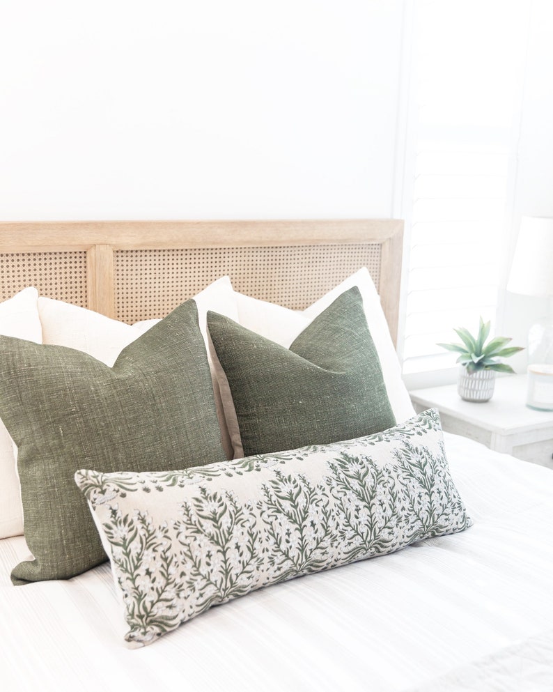 May include: A bed with a light wood headboard and white bedding. Two green square pillows and a long, rectangular pillow with a green floral pattern are arranged on the bed. A small potted plant and a lamp are on a bedside table.