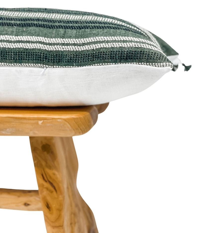 Pode incluir: Uma almofada decorativa com um padr&atilde;o listrado verde e branco na parte superior e uma parte inferior branca. A almofada est&aacute; sobre um pequeno banco de madeira clara.