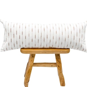 Pu&ograve; includere: Un lungo cuscino decorativo rettangolare con sfondo bianco e trattini marroni verticali. Il cuscino &egrave; appoggiato su uno sgabello di legno marrone chiaro. Le dimensioni del cuscino sono di circa 30 cm per 91 cm.