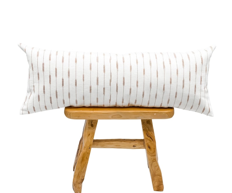 Pu&ograve; includere: Un lungo cuscino decorativo rettangolare con sfondo bianco e trattini marroni verticali. Il cuscino &egrave; appoggiato su uno sgabello di legno marrone chiaro. Le dimensioni del cuscino sono di circa 30 cm per 91 cm.