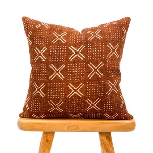 May include: A square, rust-colored decorative pillow with a repeating pattern of white crosses and dots. The pillow is placed on a small, light-colored wooden stool. The pillow has a textured appearance.