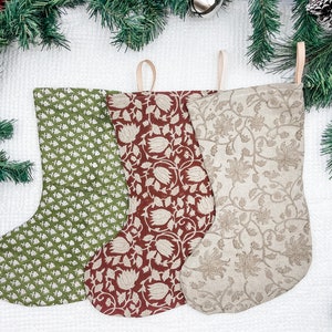 Puede incluir: Tres calcetines de Navidad con diferentes patrones florales. Los calcetines están hechos de una tela natural y tienen un aspecto rústico. Los calcetines son verdes, rojos y beige.