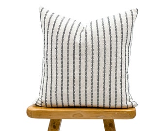 May include: A square decorative pillow with a white background and vertical black stripes. The pillow is placed on a small, light-colored wooden stool. The pillow has a textured appearance, suggesting a woven fabric.
