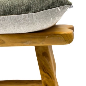 May include: A two-toned decorative pillow with a textured, olive green top and a light gray bottom. The pillow rests on a wooden stool with a natural finish, showcasing the pillow's design and texture. The pillow is a home decor item.