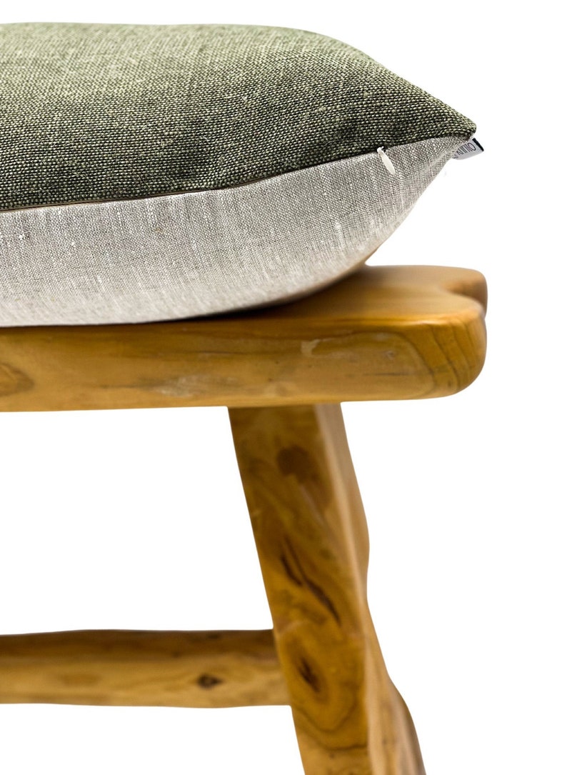 May include: A two-toned decorative pillow with a textured, olive green top and a light gray bottom. The pillow rests on a wooden stool with a natural finish, showcasing the pillow's design and texture. The pillow is a home decor item.