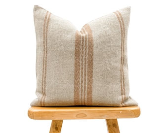 May include: A beige and brown striped throw pillow with a subtle herringbone pattern. The pillow is sitting on a wooden stool.