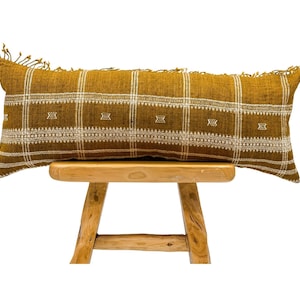 May include: A long, rectangular mustard yellow throw pillow with a white geometric pattern and fringe trim. The pillow is sitting on a wooden stool.