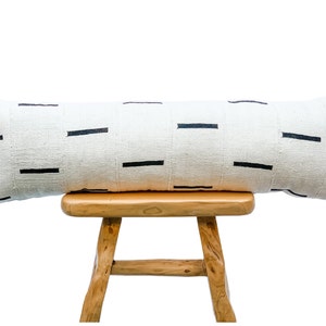 May include: A long white bolster pillow with black horizontal stripes sits on a wooden stool. The pillow is made of a natural fabric and has a simple, modern design.