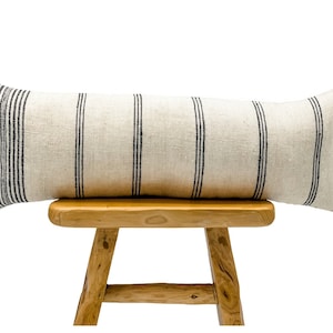 May include: A long, rectangular pillow with black and white stripes. The pillow is made of a natural linen fabric and is sitting on a wooden stool.