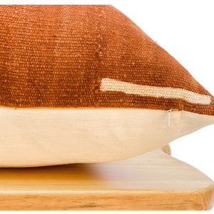 May include: A rust-colored throw pillow with a white stripe across the front. The pillow is made of a woven fabric and has a zipper closure.