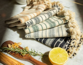 May include: A stack of five kitchen towels with fringe edges. The towels are in a variety of patterns, including stripes, checks, and geometric designs. The towels are folded and arranged on a white marble surface. A wooden rolling pin and a lemon are also in the image.