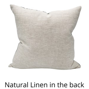 May include: A square, neutral-toned linen pillow with a textured appearance. The pillow has a dark trim and the text "Natural Linen in the back" at the bottom. The pillow is likely for home decor.
