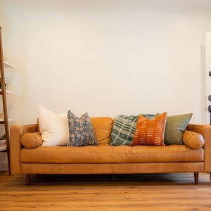 May include: A brown leather sofa with several decorative pillows in various colors and patterns. The pillows include white, blue floral, green plaid, orange geometric, and olive green. A wooden ladder shelf and a woven basket are also visible.