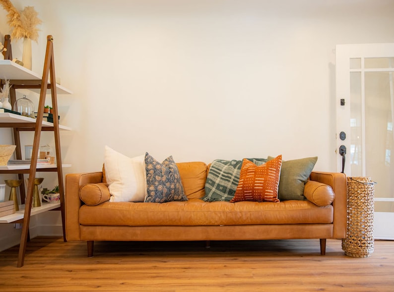 May include: A brown leather sofa with several decorative pillows in various colors and patterns. The pillows include white, blue floral, green plaid, orange geometric, and olive green. A wooden ladder shelf and a woven basket are also visible.