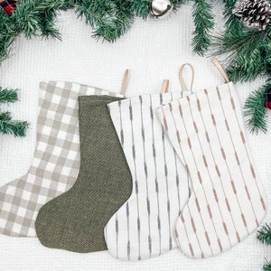 Puede incluir: Cuatro calcetines de Navidad con diferentes patrones. Uno es de cuadros marrones y blancos, otro es de color verde oscuro sólido, uno es de rayas blancas y grises y otro es de rayas blancas y marrones.