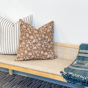 May include: A wooden bench with a beige cushion and a blue throw blanket. There are three pillows on the bench, one with a brown floral pattern and two with white and black stripes.
