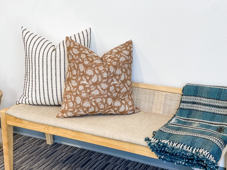 May include: A wooden bench with a beige cushion and a blue throw blanket. There are three pillows on the bench, one with a brown floral pattern and two with white and black stripes.