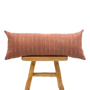 Puede incluir: Un cojín lumbar largo y rectangular de tela de lino color óxido con líneas blancas bordadas. El cojín está sentado en un taburete de madera.