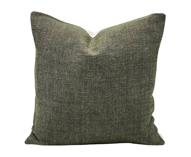 May include: Square decorative pillow with a textured, olive green fabric cover. The pillow has a neutral-colored backing and a visible seam. The fabric has a woven appearance, adding visual interest to the design. The pillow is suitable for home decor.