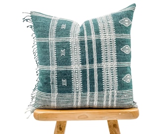 May include: A teal blue and white patterned throw pillow with fringe on the edges. The pillow is sitting on a wooden stool.