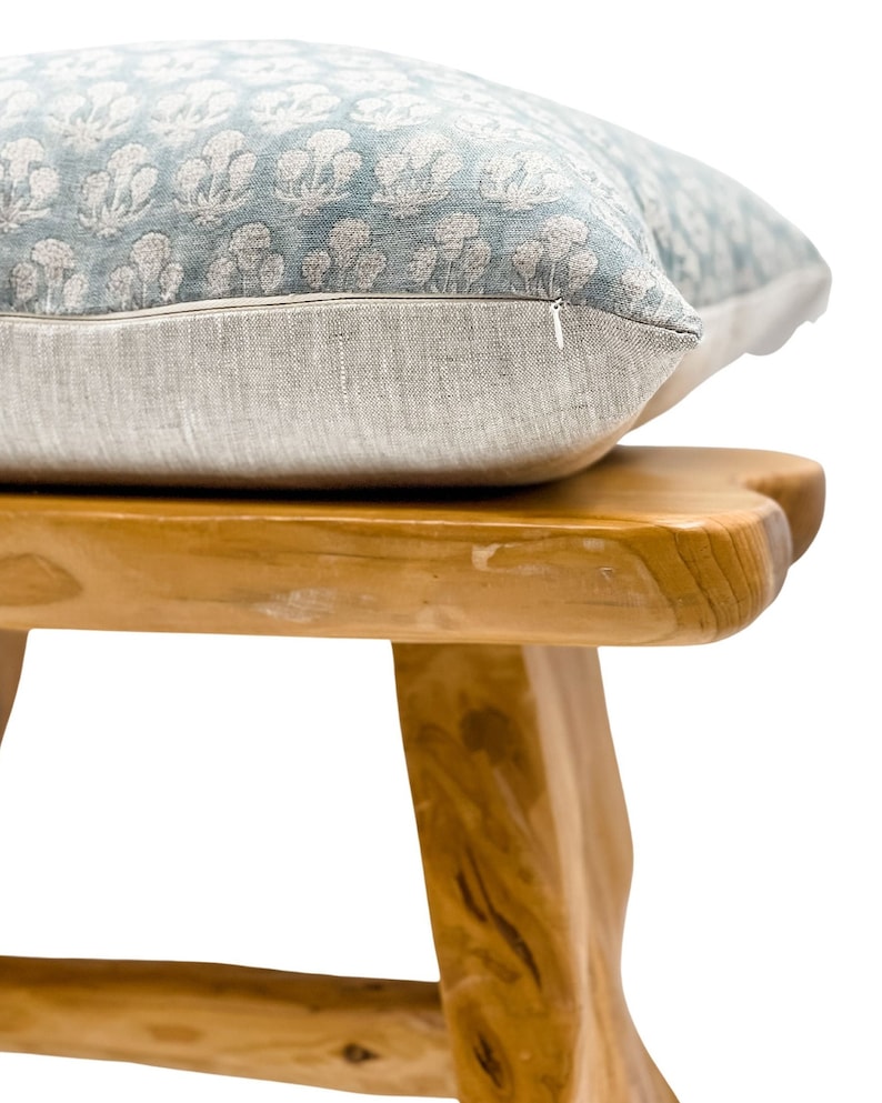 May include: A light blue floral patterned pillow with a zipper closure sits on top of a wooden stool with a natural finish.