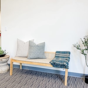 May include: A wooden bench with a light gray cushion and two pillows. One pillow has a black and white striped pattern, and the other has a blue and white geometric pattern. A blue and white throw blanket is draped over the bench. A small potted olive tree is on the right side of the image.