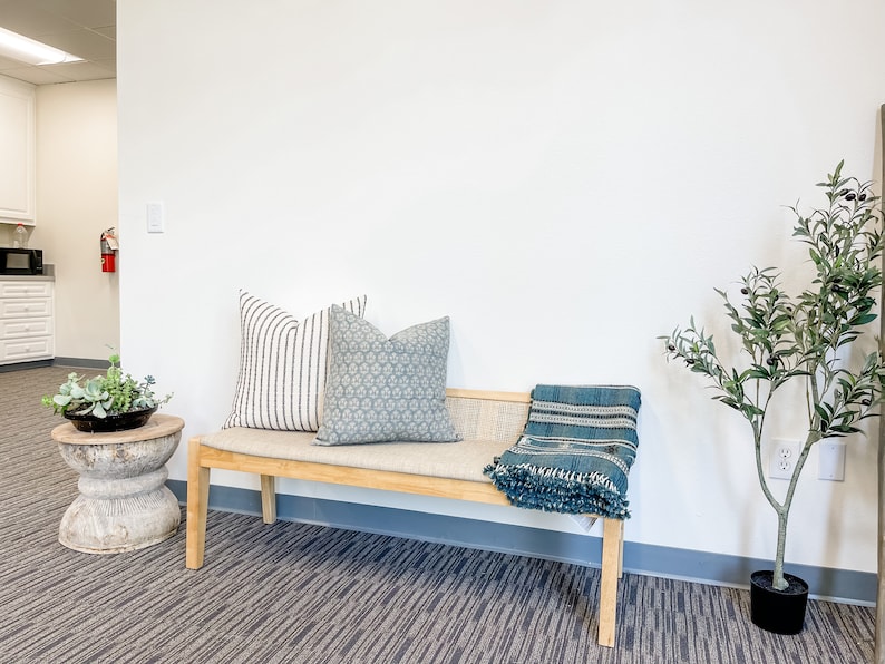 May include: A wooden bench with a light gray cushion and two pillows. One pillow has a black and white striped pattern, and the other has a blue and white geometric pattern. A blue and white throw blanket is draped over the bench. A small potted olive tree is on the right side of the image.