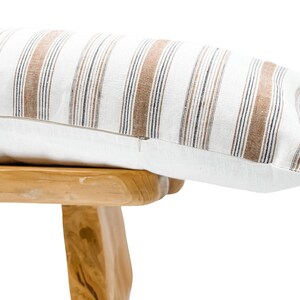 Pu&ograve; includere: Un cuscino decorativo rettangolare con base bianca e strisce verticali marroni e blu navy. Il cuscino ha una chiusura a cerniera e poggia su uno sgabello di legno chiaro.