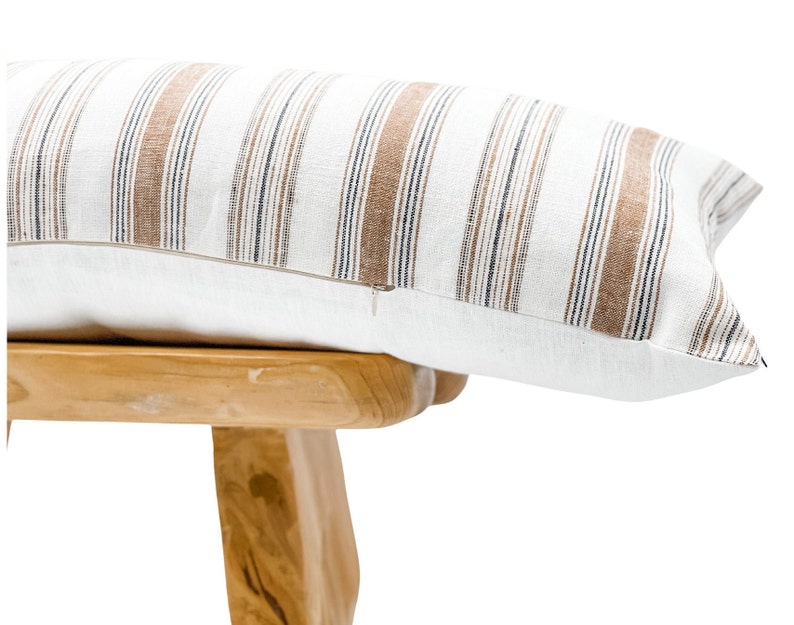 Pu&ograve; includere: Un cuscino decorativo rettangolare con base bianca e strisce verticali marroni e blu navy. Il cuscino ha una chiusura a cerniera e poggia su uno sgabello di legno chiaro.