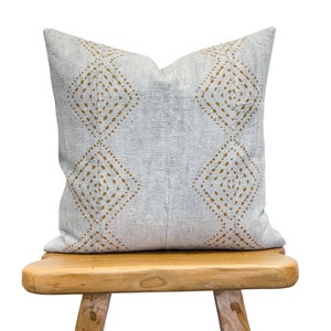 May include: A gray throw pillow with a geometric pattern in gold. The pillow is sitting on a wooden stool.