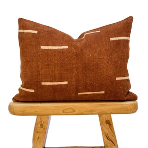 May include: A rectangular cushion in a rust colour, featuring horizontal cream stripes. The cushion is made of textured fabric and rests on a small, light-coloured wooden stool.